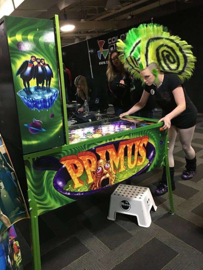 Woman playing pinball at Comic-Con with vibrant green and yellow costume headpiece.