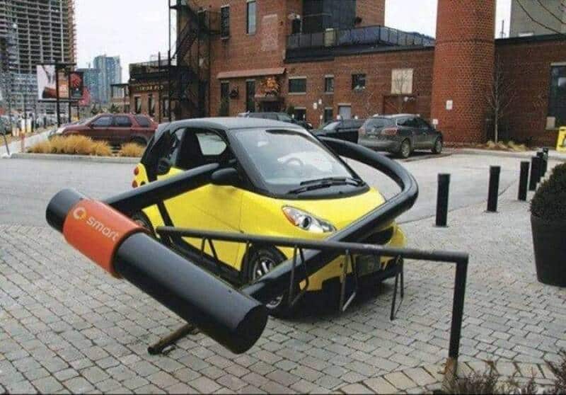 Small yellow electric car trapped under oversized smartphone sculpture in urban setting.