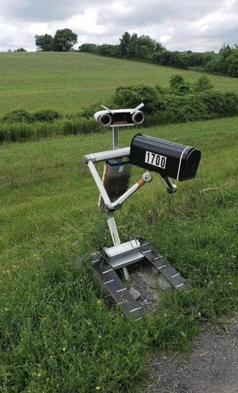 Robot sculpture in a grassy field with rolling hills, trees, and a cloudy sky.