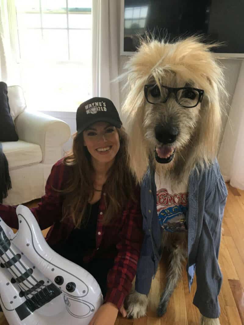 Playful dog dressed as a rock star with glasses and a woman smiling in a cozy living room.