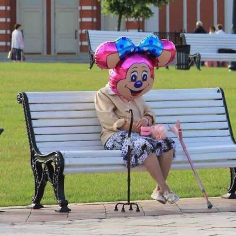 Minnie Mouse costume worn by elderly woman sitting on park bench, smiling and enjoying the day.
