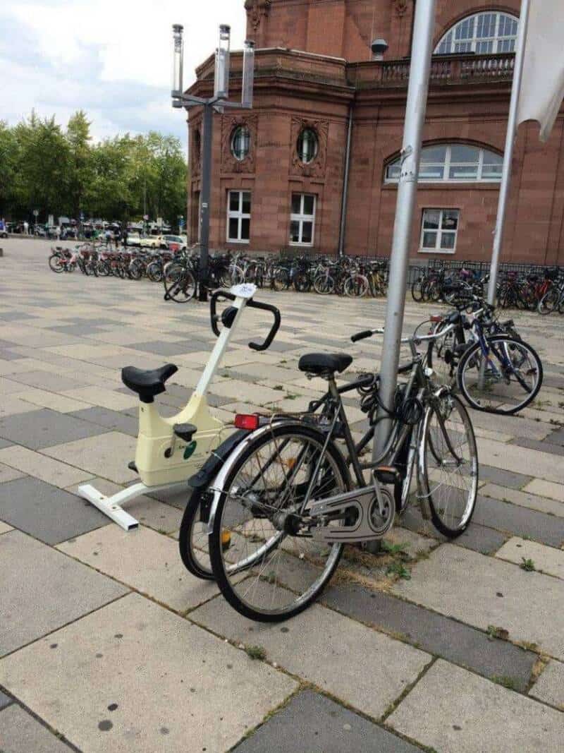 Bicycle locked to a pole with a stationary exercise bike nearby in an urban setting.