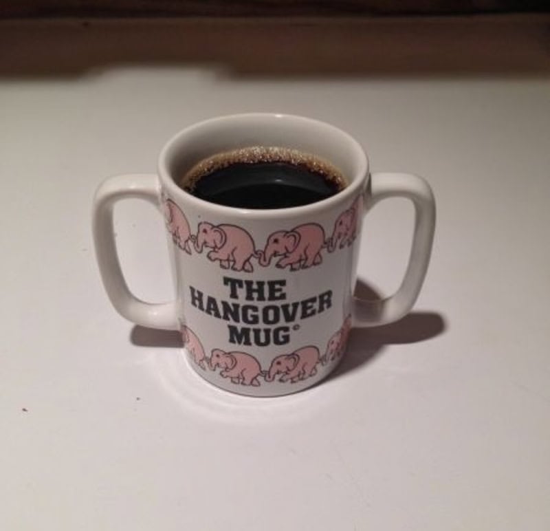 Coffee mug with "The Hangover Mug" text and pink elephant pattern, filled with black coffee.