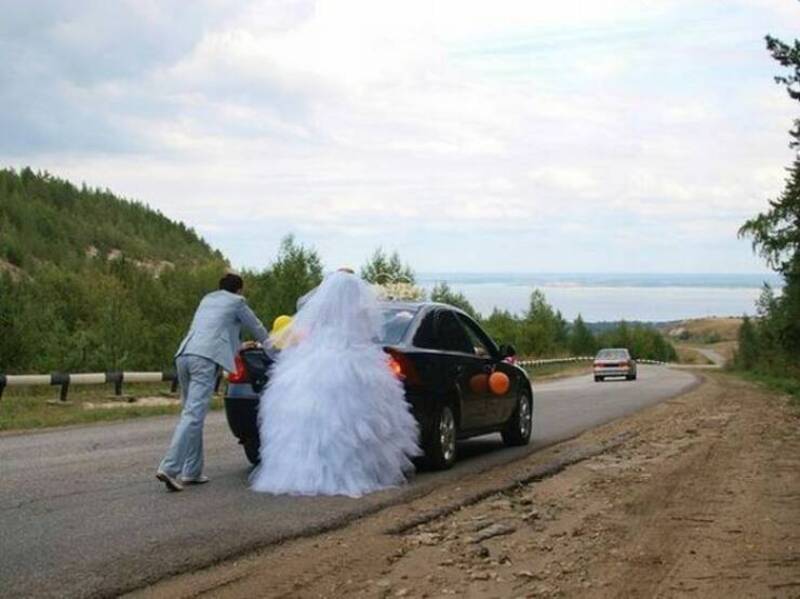 Bride in wedding dress and groom pushing car on rural road with scenic background.