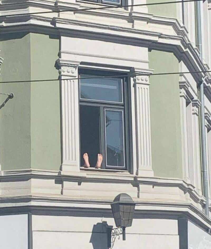 View of a person with legs hanging out of a second-story window of a green building.