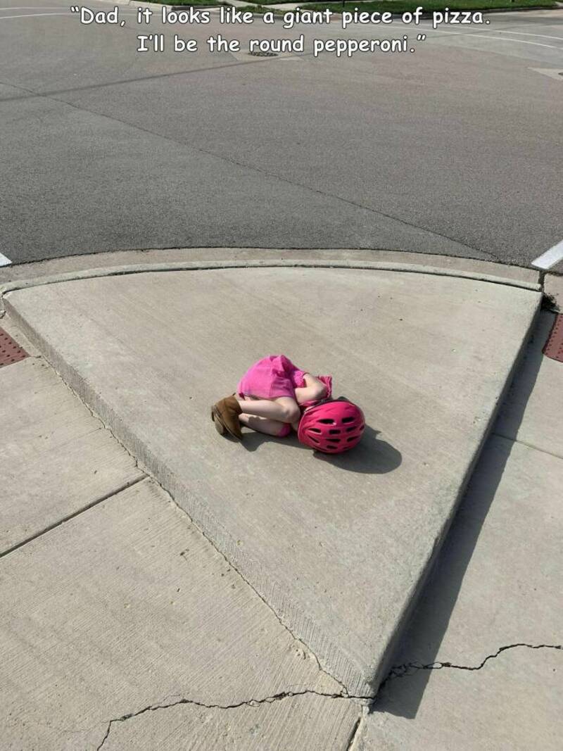 Child lying on sidewalk with pink helmet, pretending a pizza slice is a giant piece of pizza.