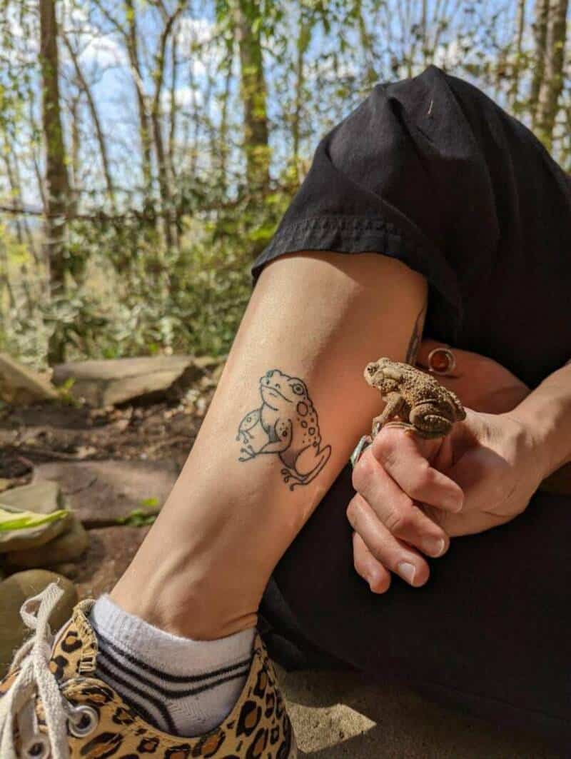 Person holding a small lizard with a tattoo on their arm outdoors.