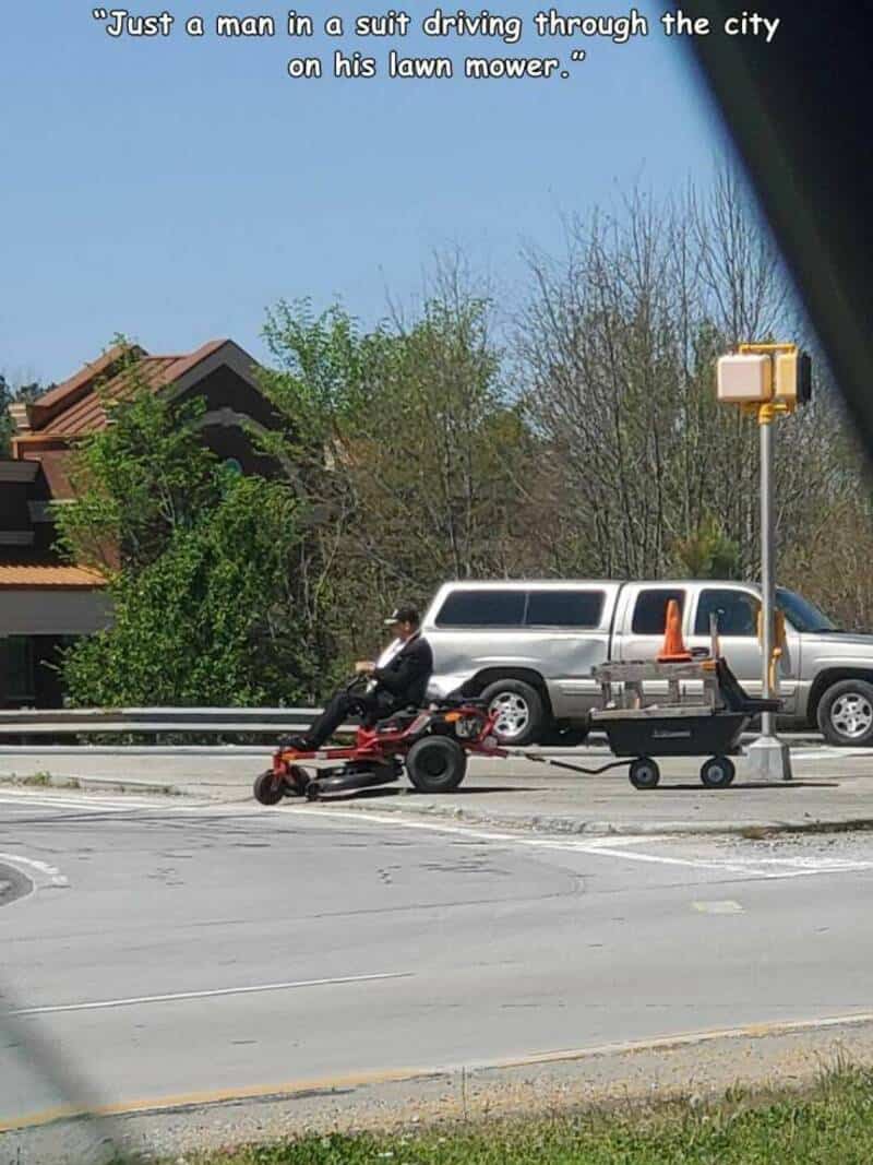 Man in formal attire riding a lawn mower pulling a cart on a busy road.