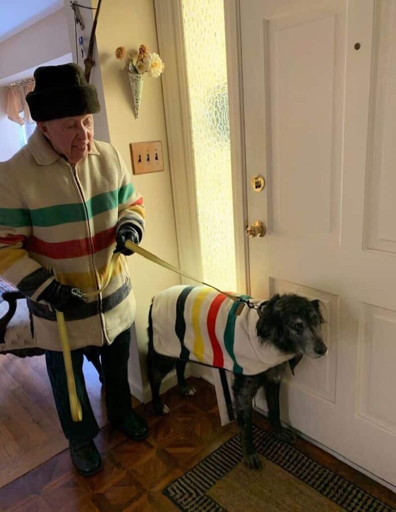 Elderly woman in colorful sweater with dog wearing matching sweater at door.