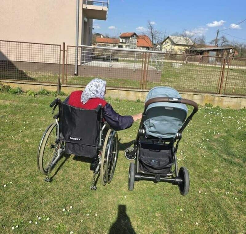 Wheelchair and stroller outdoors on a sunny day, with a fence and residential buildings behind.