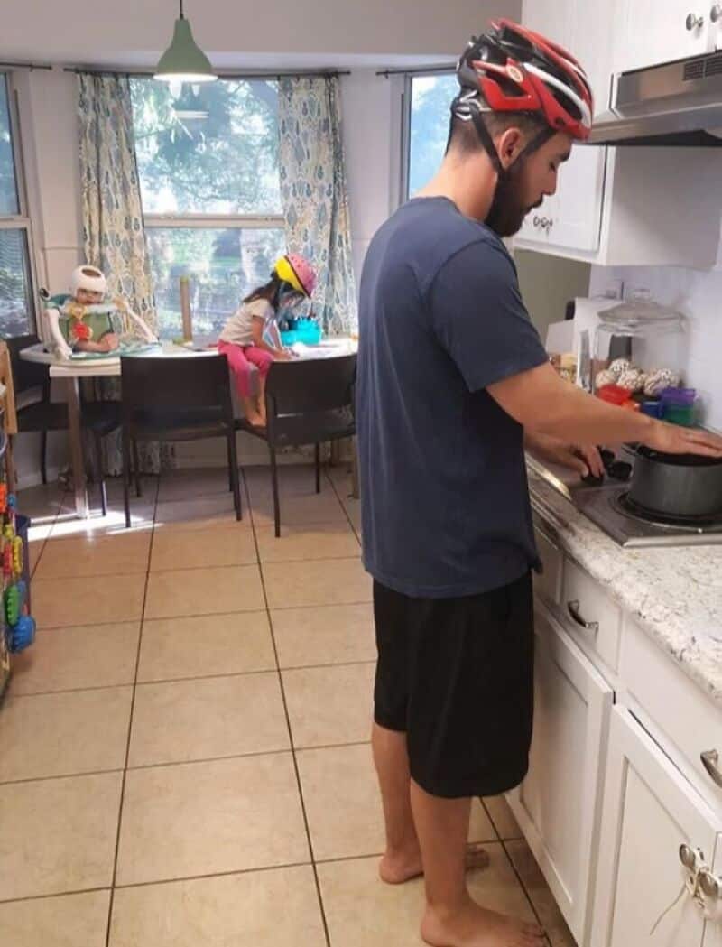 Man cooking in kitchen while children play at the table in the background.