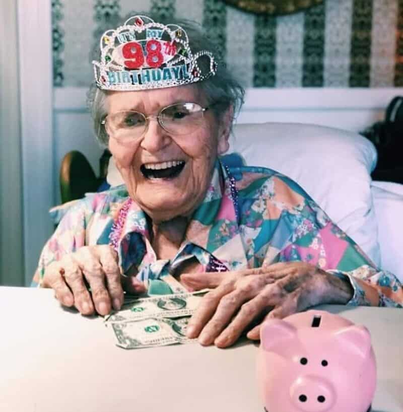 Elderly woman celebrating 98th birthday with money and pink piggy bank.