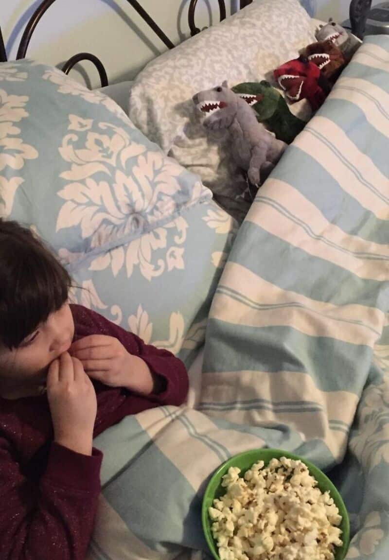 Child watching plush dinosaur toys in bed with popcorn bowl.