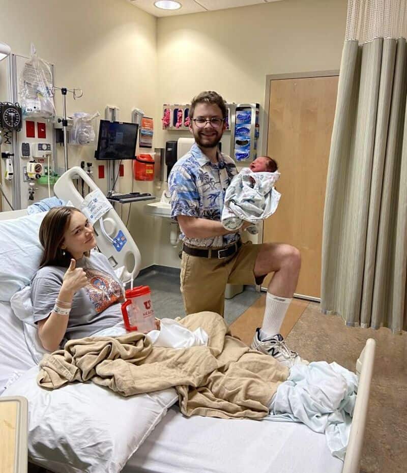 Happy father holding newborn baby in hospital room with mother resting in bed.