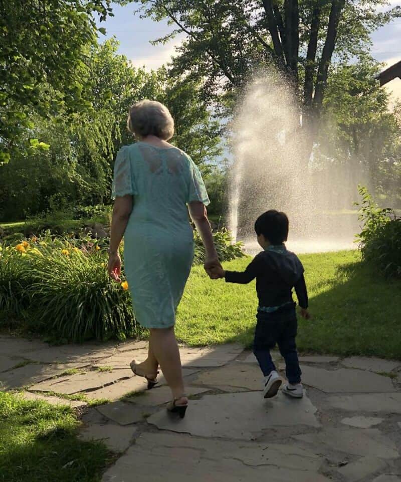 Mother and child walking hand in hand in a lush garden with a water fountain in the background.