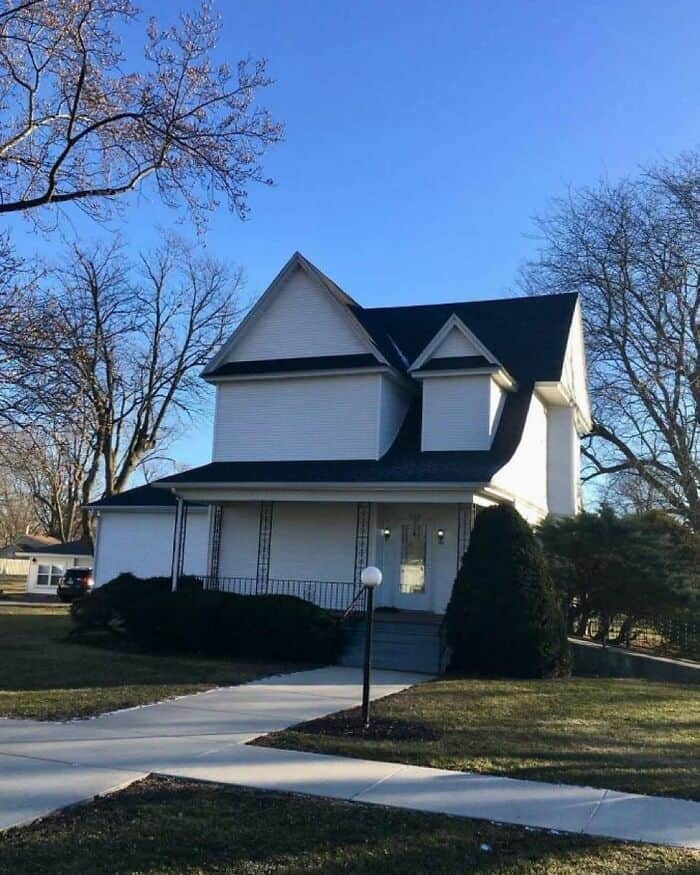 Victorian-style white house with black roof and front porch, surrounded by trees and a clear blue sk.