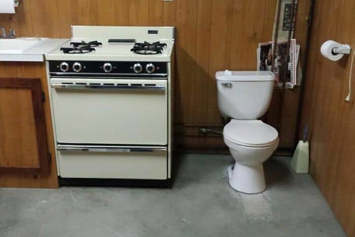 Small kitchen with stove and toilet in a wooden-paneled room.