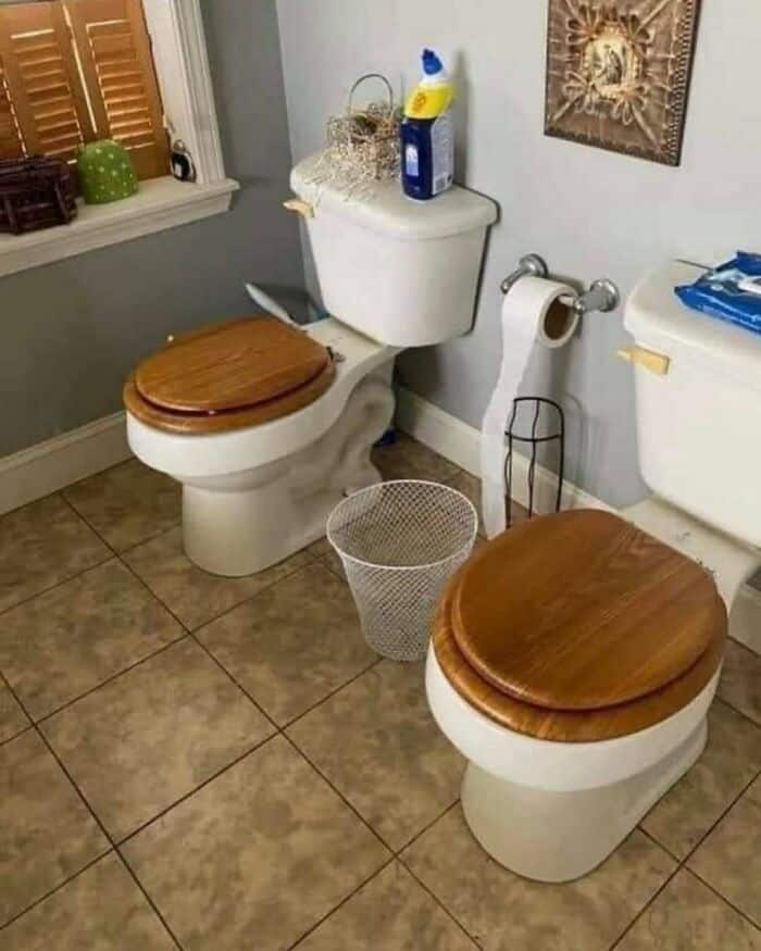 Toilet with two wooden seats, cleaning supplies on the tank, and a wastebasket on the tiled floor.