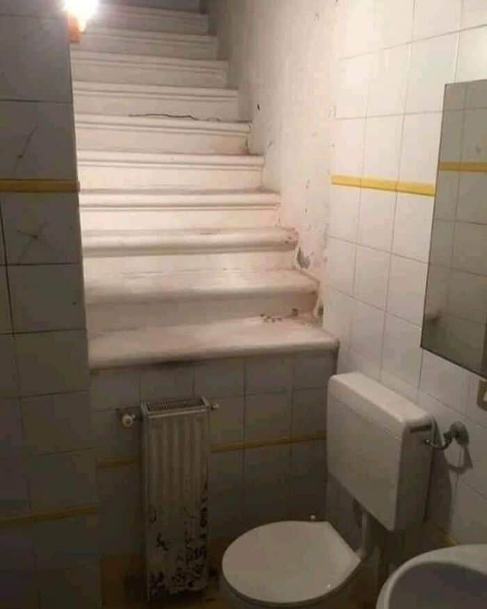 Stairs above a bathroom with a radiator and toilet in a tiled room.