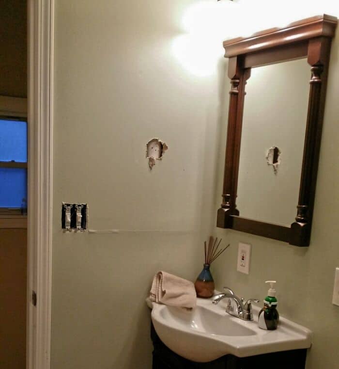 Damaged bathroom wall with holes and incomplete repairs, featuring a mirror and sink. A humorous exa.