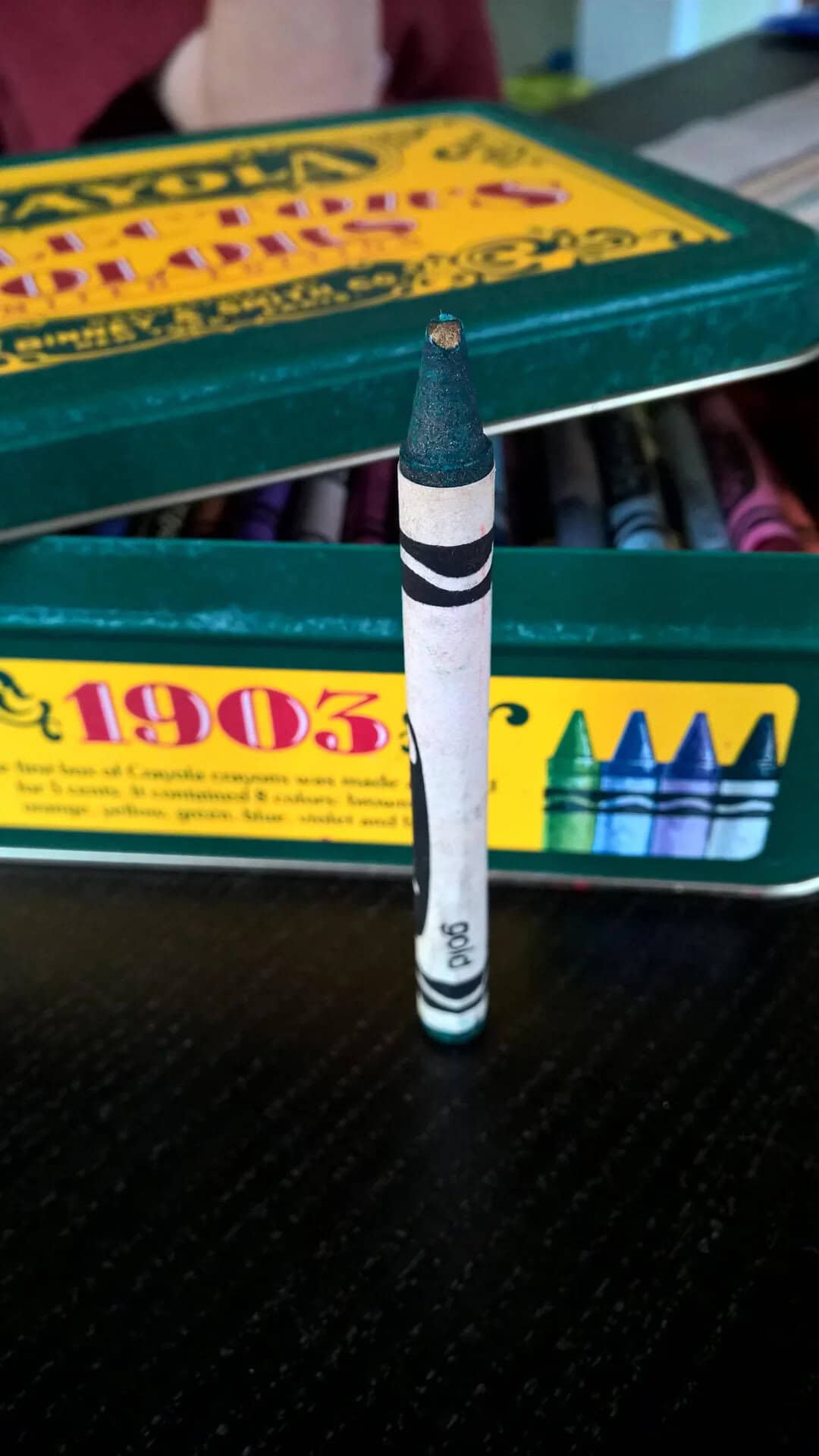 Crayon standing upright in front of a box of Crayola crayons on a black surface.