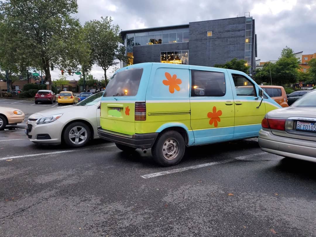 Vintage van with colorful flower decals, parked among modern cars in an urban parking lot. The van f.