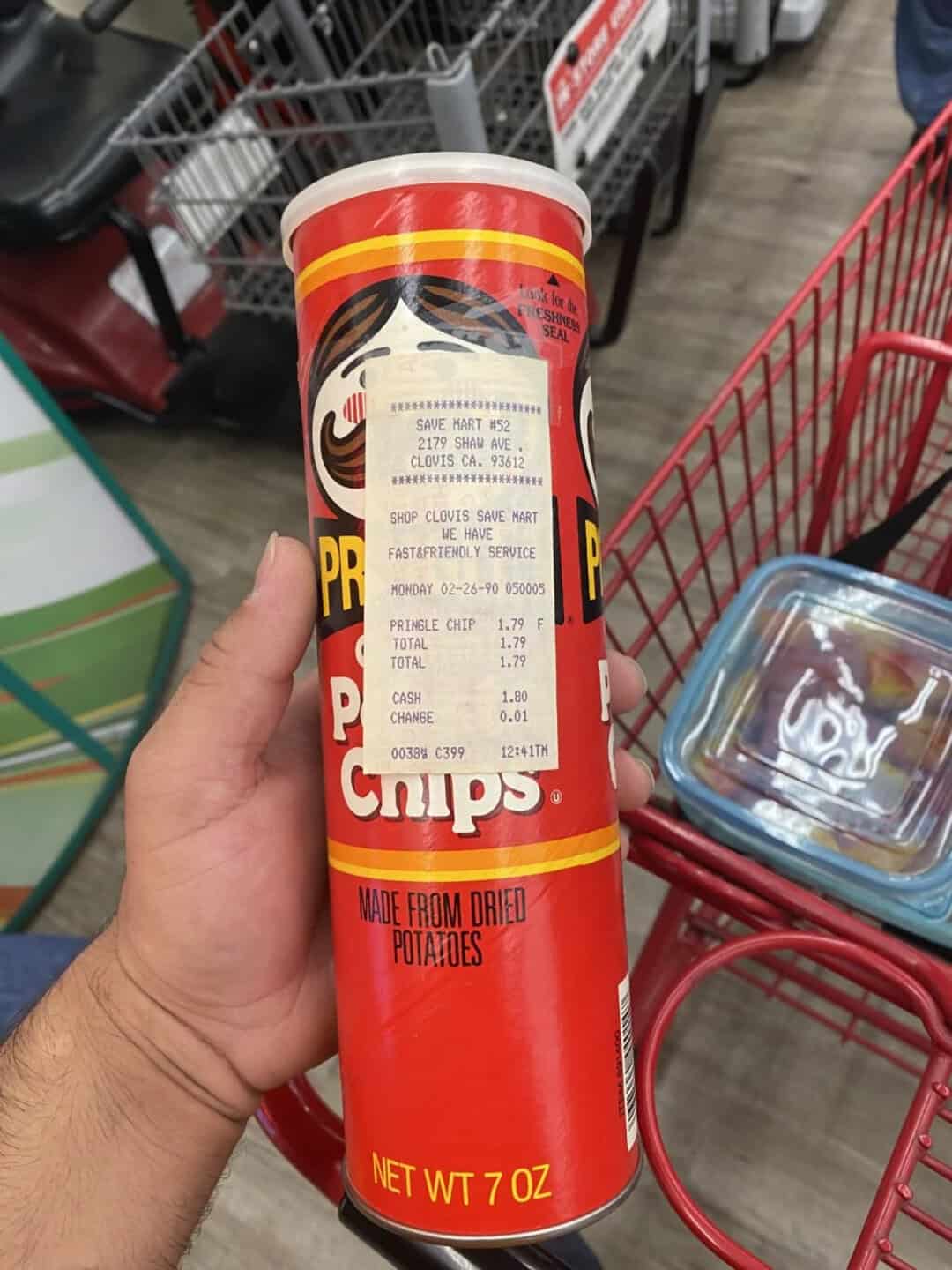Hand holding a Pringles can with a grocery store shopping cart in the background.