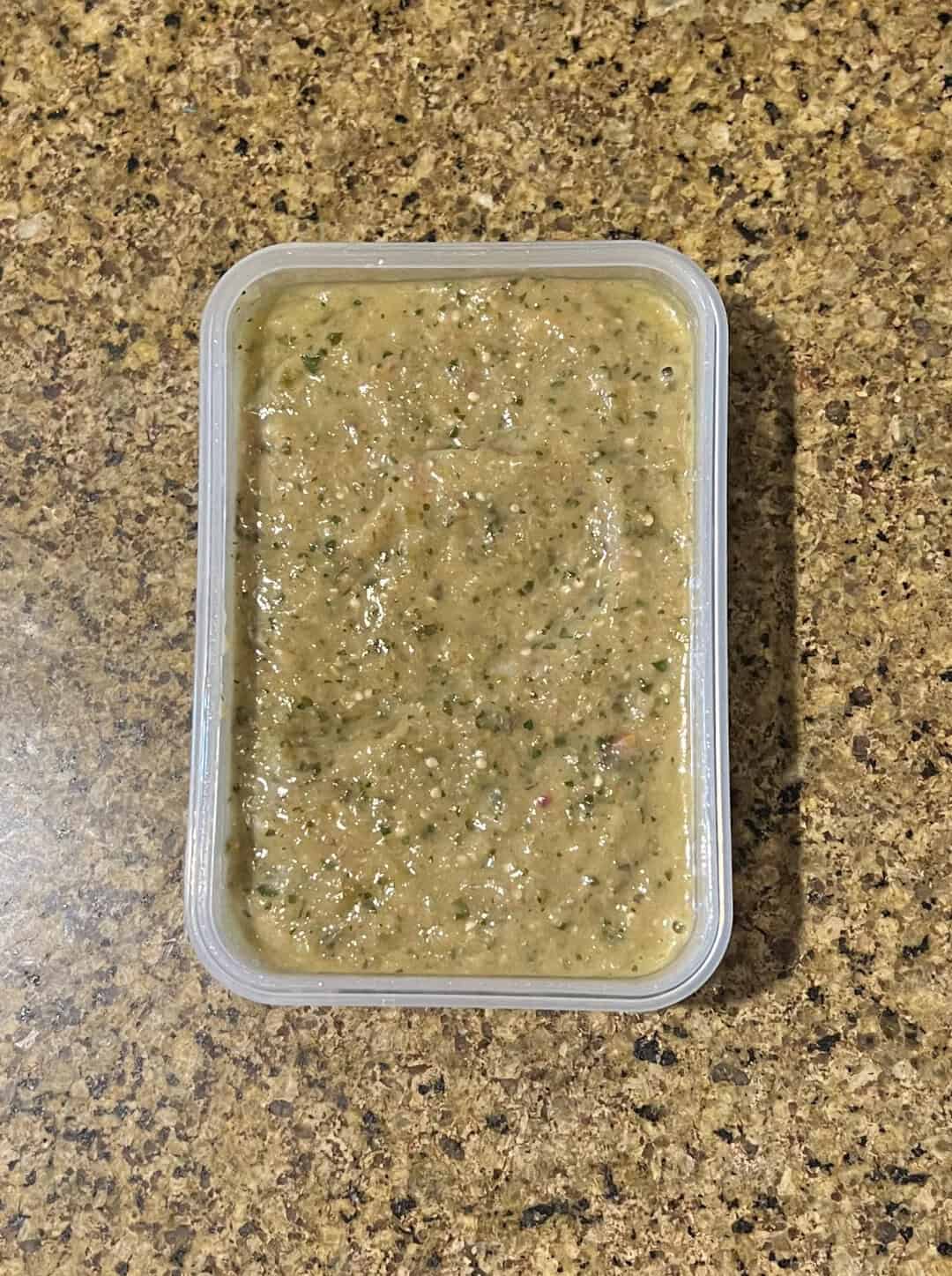 Guacamole dip with herbs in a clear plastic container on a speckled countertop.