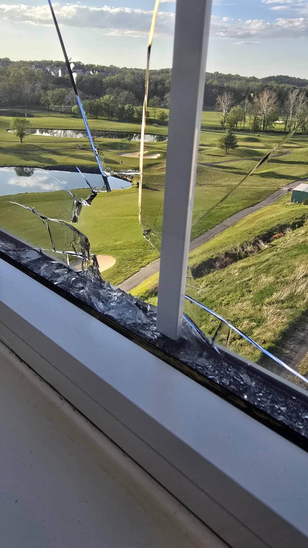 Broken window with shattered glass overlooking a golf course and pond.