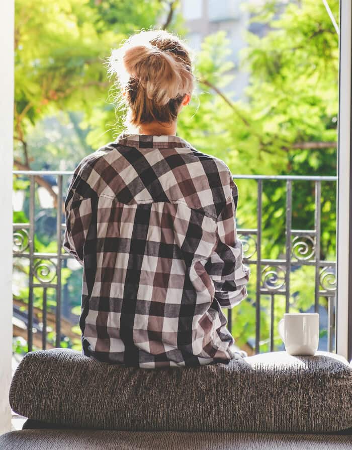 Woman sitting by window with greenery outside, wearing a checkered shirt.