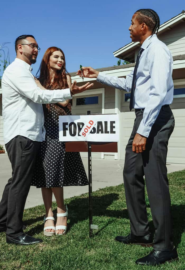 Real estate agent handing over house keys to new homeowners outside a for-sale sign.