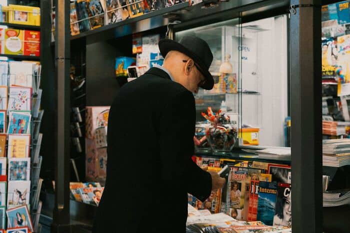 Man browsing magazines at newsstand, wearing black hat and coat.