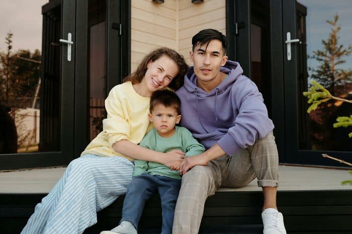 Happy family sitting on porch steps, smiling at camera, casual clothing, autumn background.