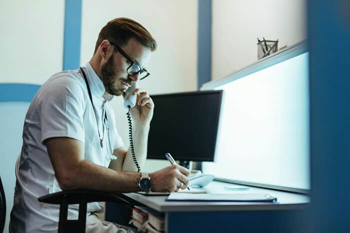 Medical professional talking on the phone in a clinical setting, taking notes at desk with computer.