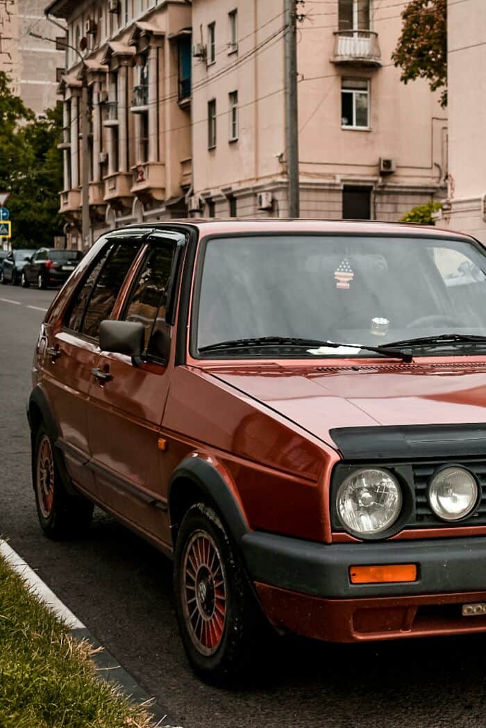 Vintage red compact hatchback parked on city street with apartment buildings in the background.