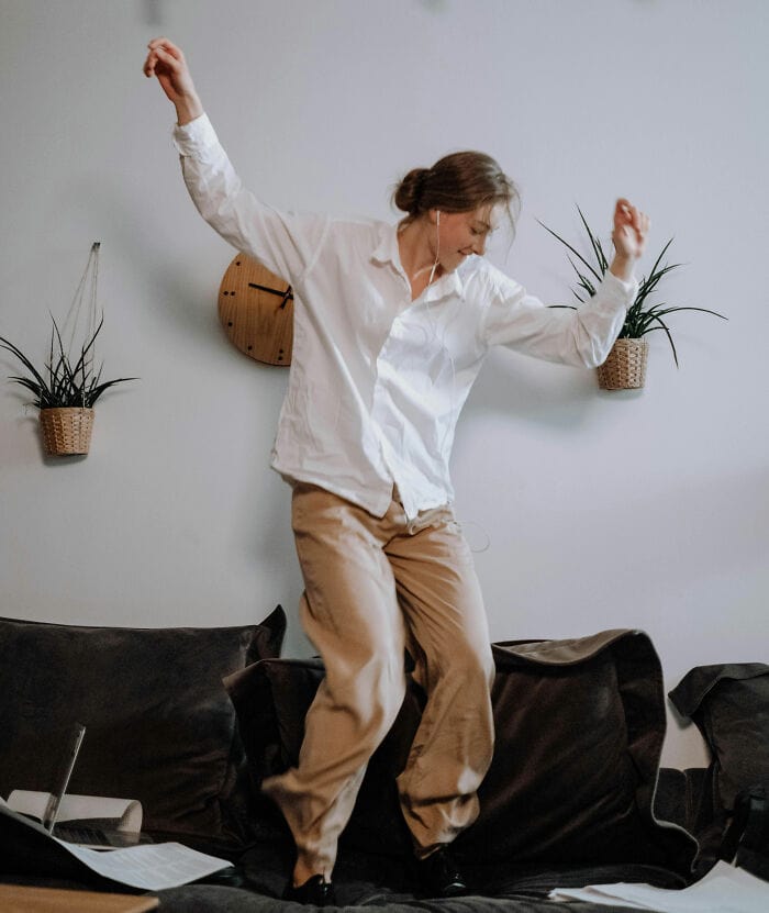 Woman dancing on sofa with arms raised in a relaxed home setting.