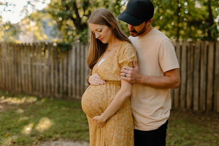 Pregnant woman with her partner in a garden, holding her belly and sharing a tender moment outdoors.