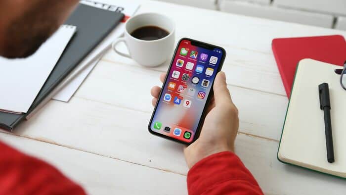 Close-up of a person holding an iPhone with app icons visible, on a white desk with coffee, notebook.
