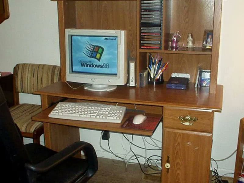 Computer desk with Windows 98 monitor, keyboard, mouse, and office supplies in a home office setting.