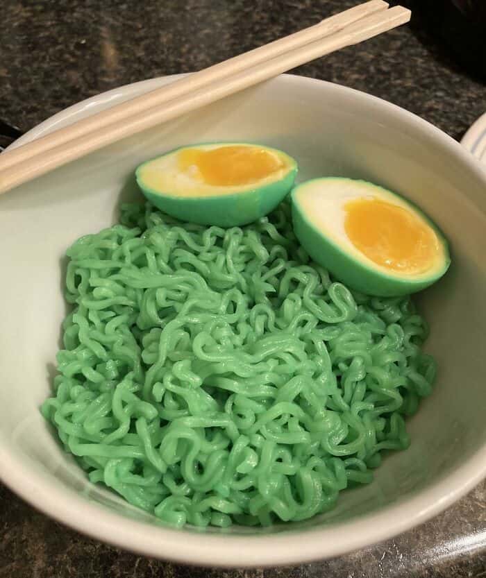 Green ramen noodles with halved soft-boiled eggs in a white bowl, on a dark countertop.