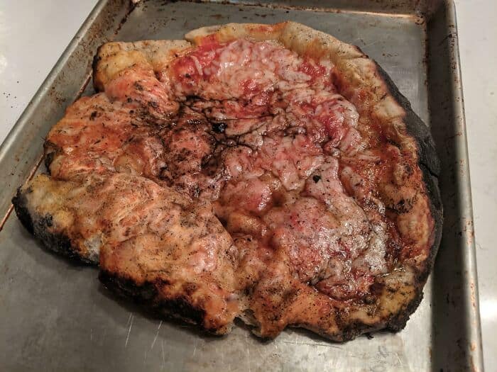 Uncooked burnt pizza with charred edges, melted cheese, and uneven toppings on a baking sheet.
