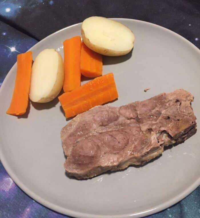 Unappealing image of a raw, overcooked piece of meat with peeled potatoes and carrots on a plate.