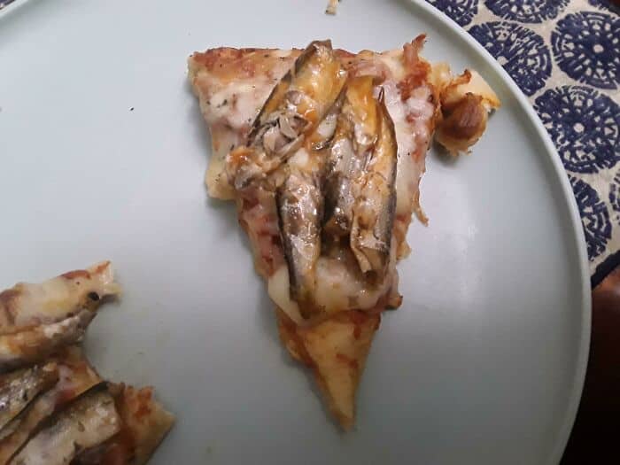 Pizza slice with small fish and melted cheese, on a white plate.