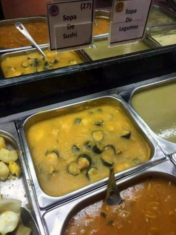 Unappetizing yellow soup with strange black and green ingredients in a cafeteria tray.