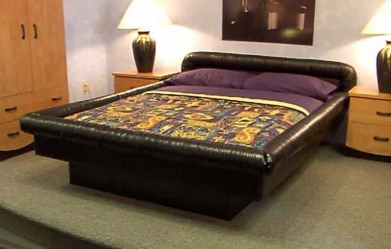 A bed with a black leather frame, purple pillows, and a colorful quilt in a bedroom.
