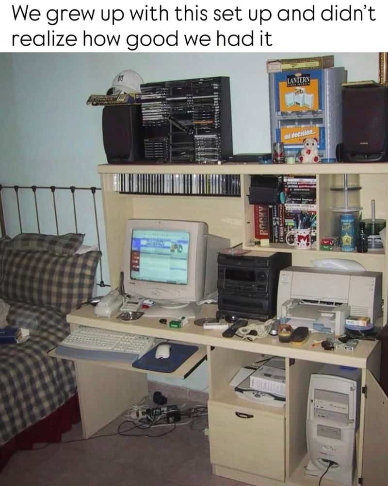 Old computer setup with a monitor, keyboard, and various electronic devices on a desk, with a bed ne.