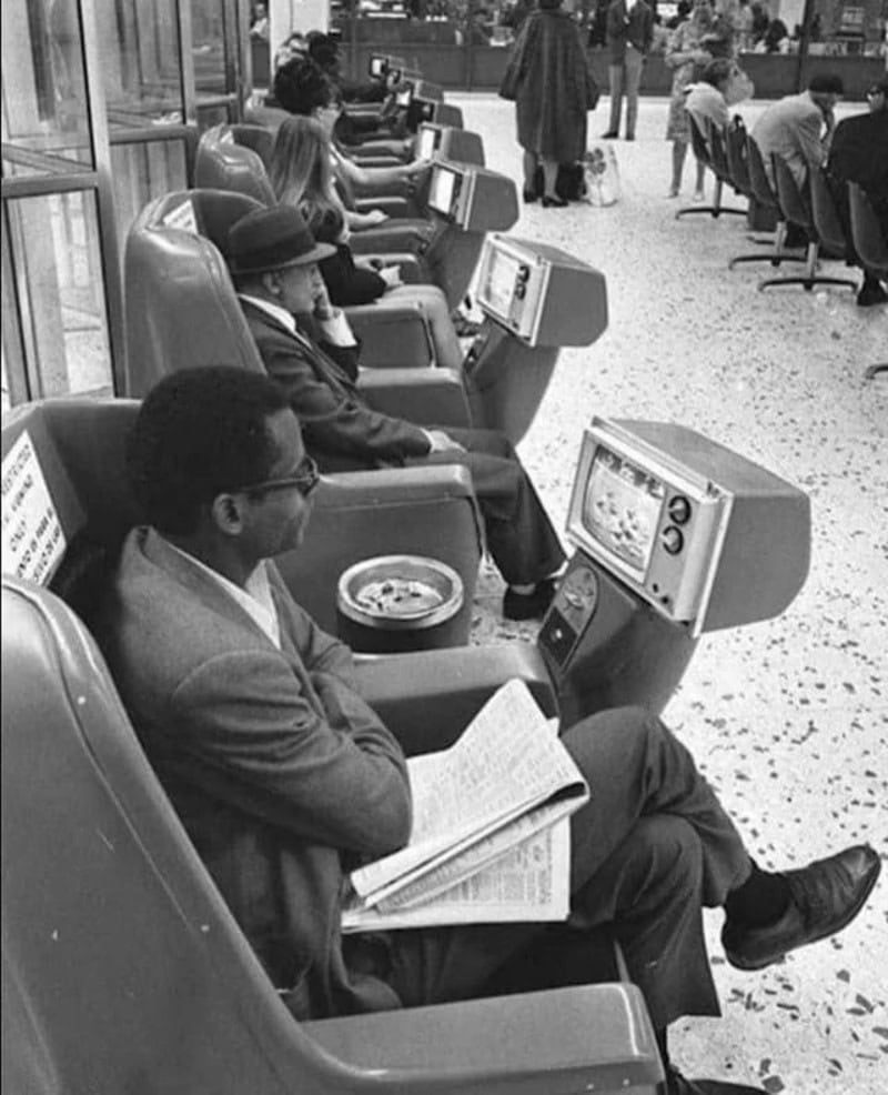 A woman reading a newspaper while waiting at an airport terminal with people using computers.