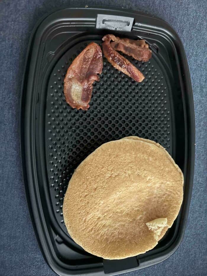 Bacon and pancake on black tray with a small piece of pancake missing.