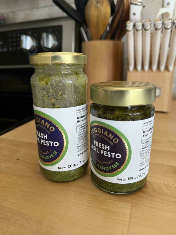 Creamy green pesto in glass jars on a kitchen countertop with knives and utensils in the background.