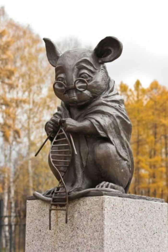 A playful mouse sculpture wearing glasses and holding a DNA strand on a stone pedestal.